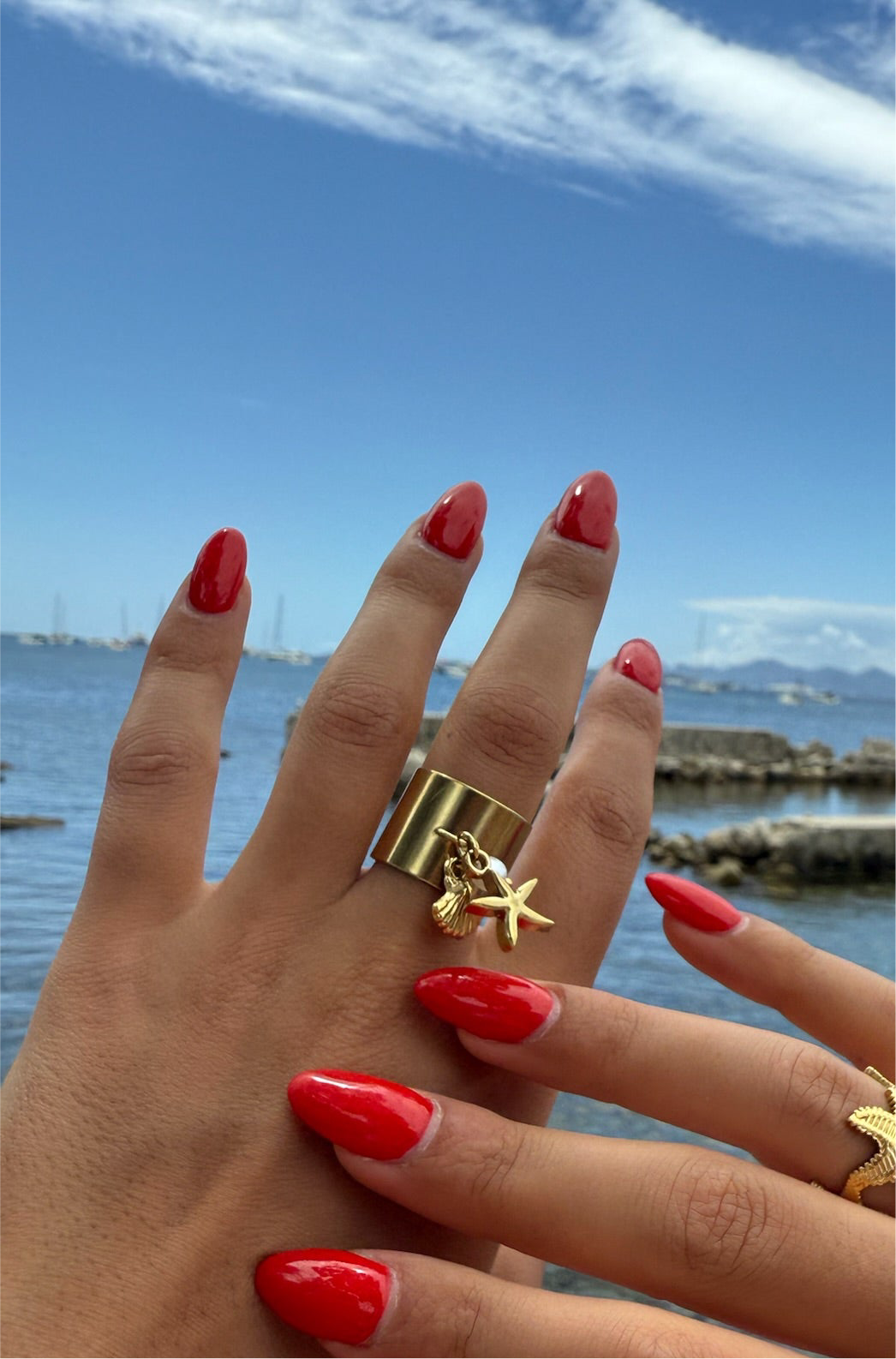 Bague dorée Armeria en acier inoxydable avec pendentifs coquillage et étoile de mer, portée avec une manucure rouge sur fond marin, signée Onaïa Bijoux.