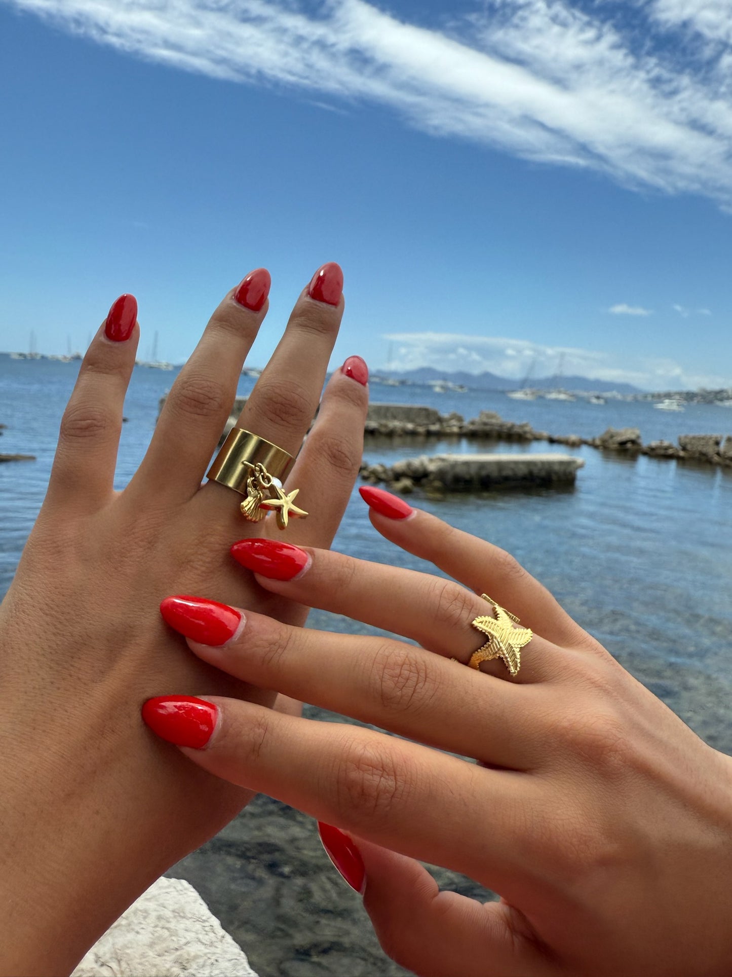 Bague dorée Schiuma d’Oro en forme d’étoile de mer signée Onaïa Bijoux, portée au bord de l’eau avec une manucure rouge éclatante.