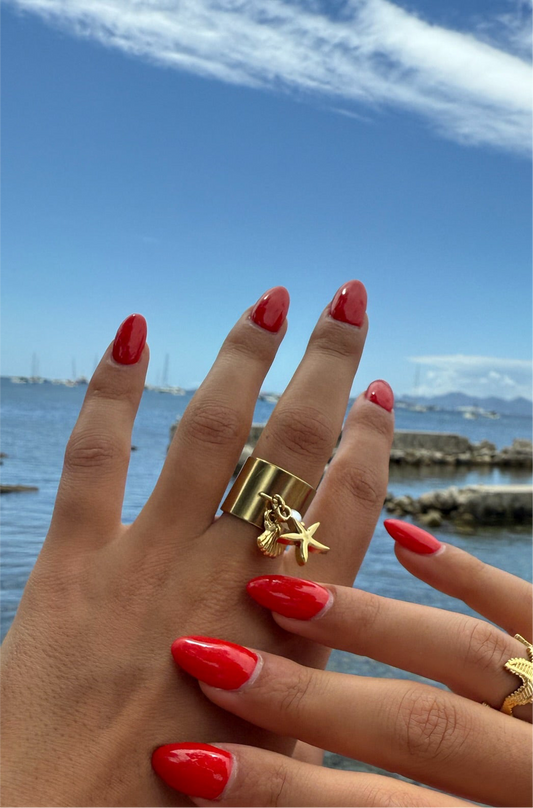 Bague dorée Armeria en acier inoxydable avec pendentifs coquillage et étoile de mer, portée avec une manucure rouge sur fond marin, signée Onaïa Bijoux.