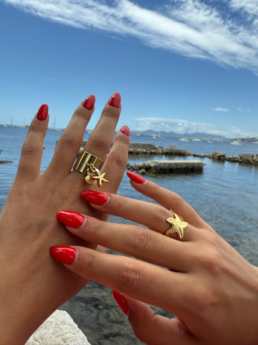 Bague dorée Schiuma d’Oro en forme d’étoile de mer signée Onaïa Bijoux, portée au bord de l’eau avec une manucure rouge éclatante.